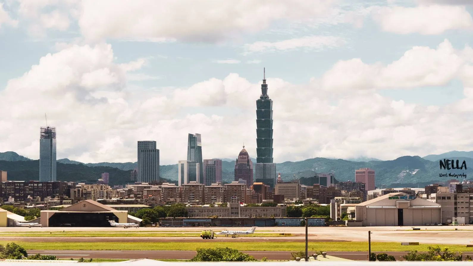 遠景中高樓天際線與山脈相接,前方是道路和低矮屋頂,中央矗立一棟細長摩天大樓