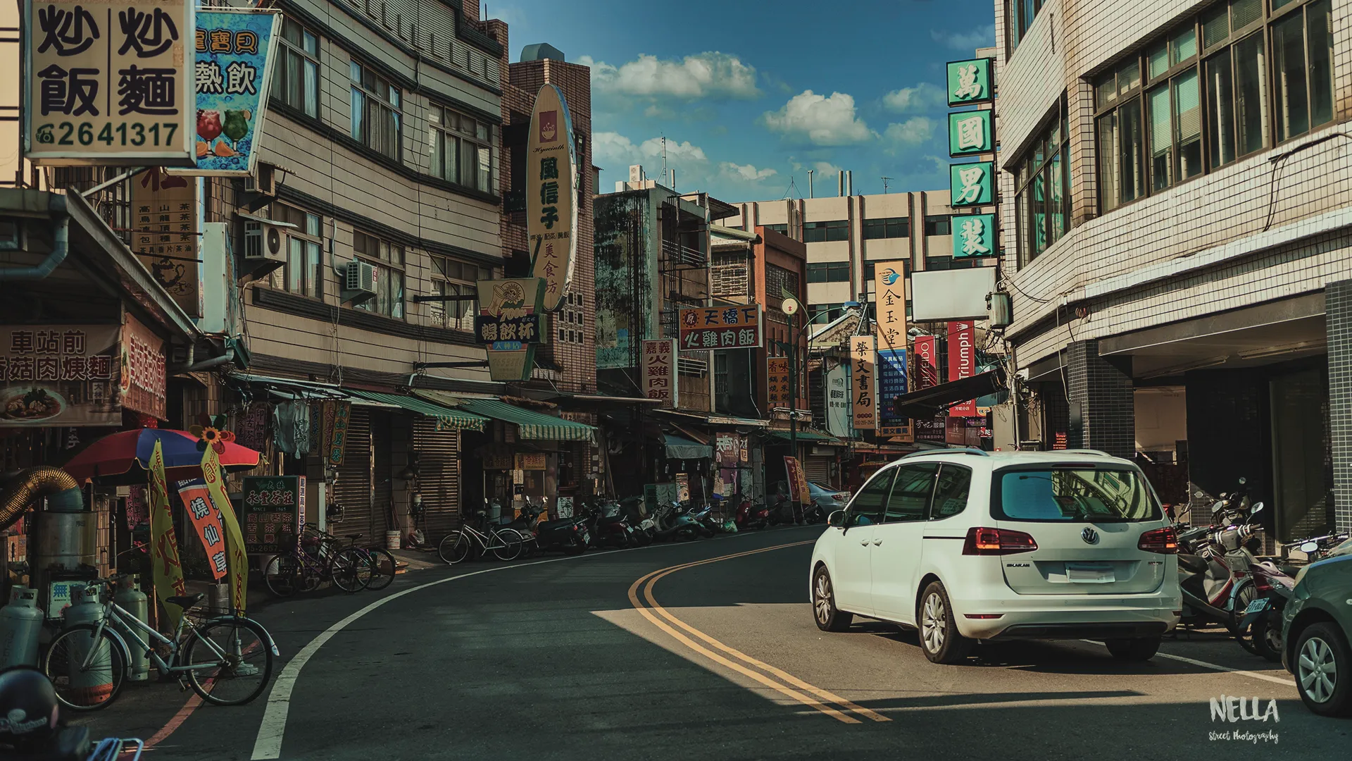 彎曲道路穿過兩側店屋,白色汽車停在右側,晴空下可見招牌、行人與機車