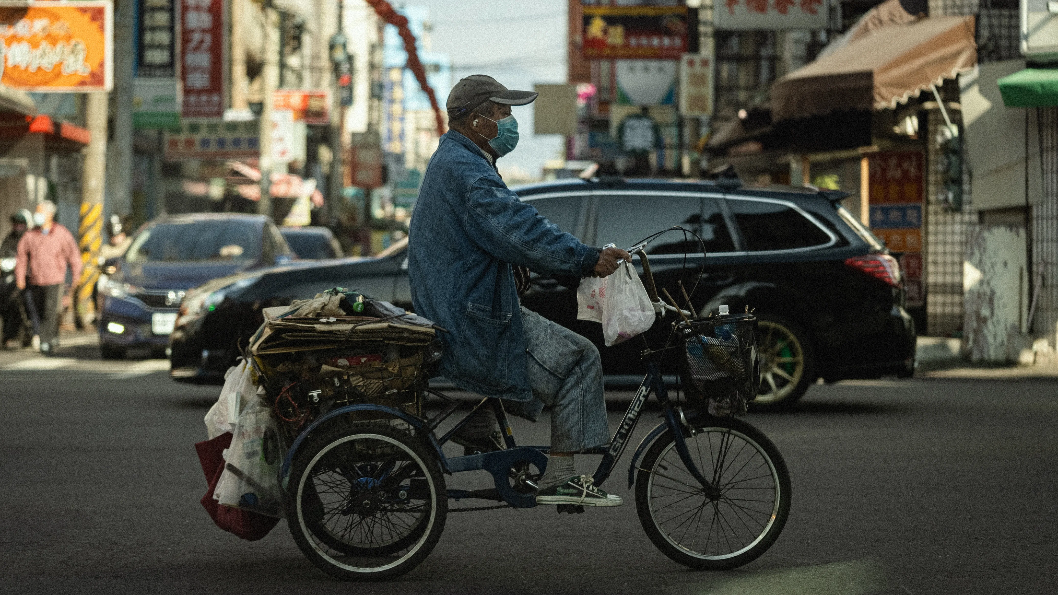 一名戴口罩的男子騎著載滿物品的三輪車穿過繁忙街道,後方有汽車與店家