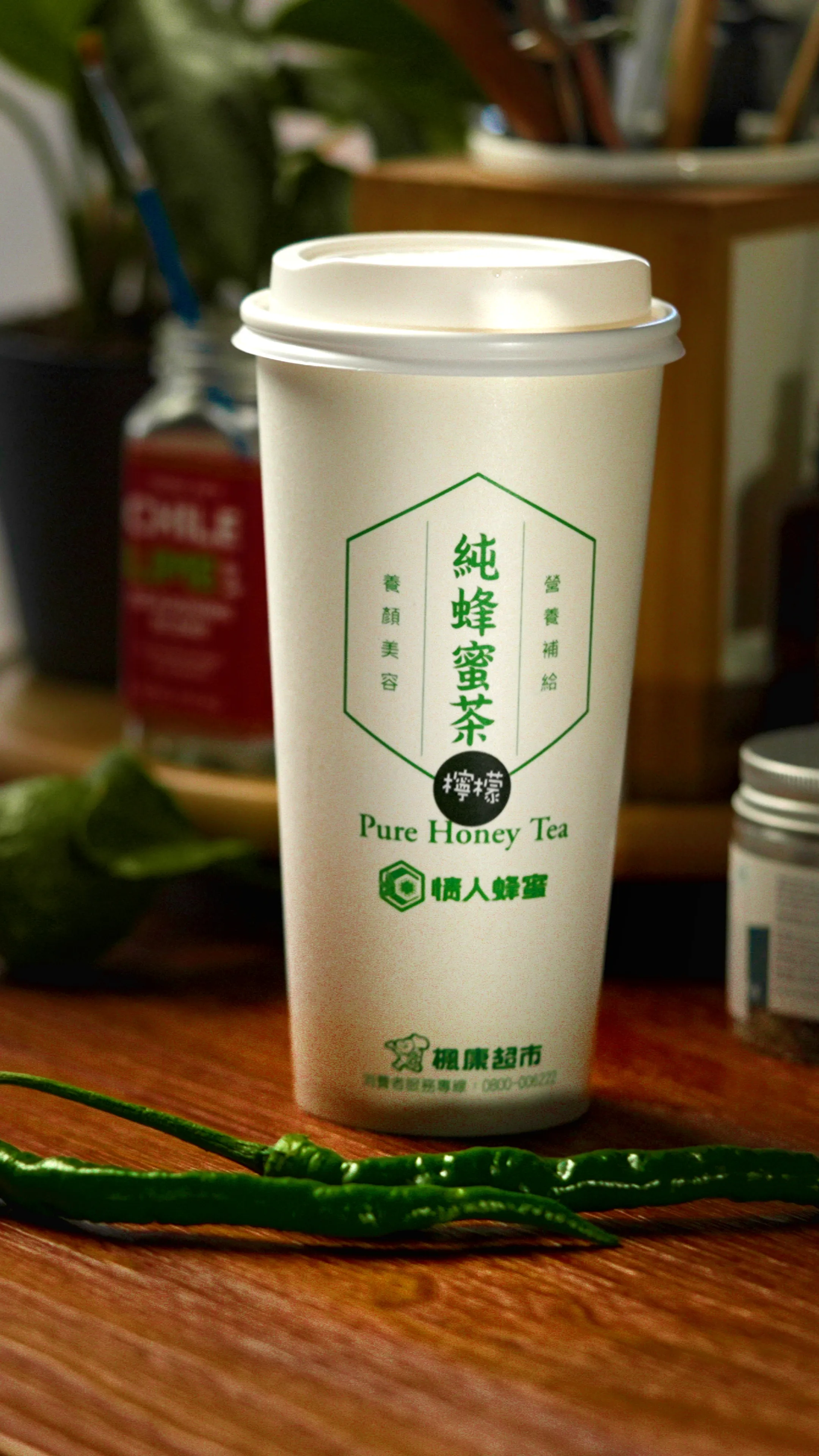 白色外帶杯立在木桌上,杯身印有純蜂蜜茶字樣,前方橫放兩根綠辣椒
